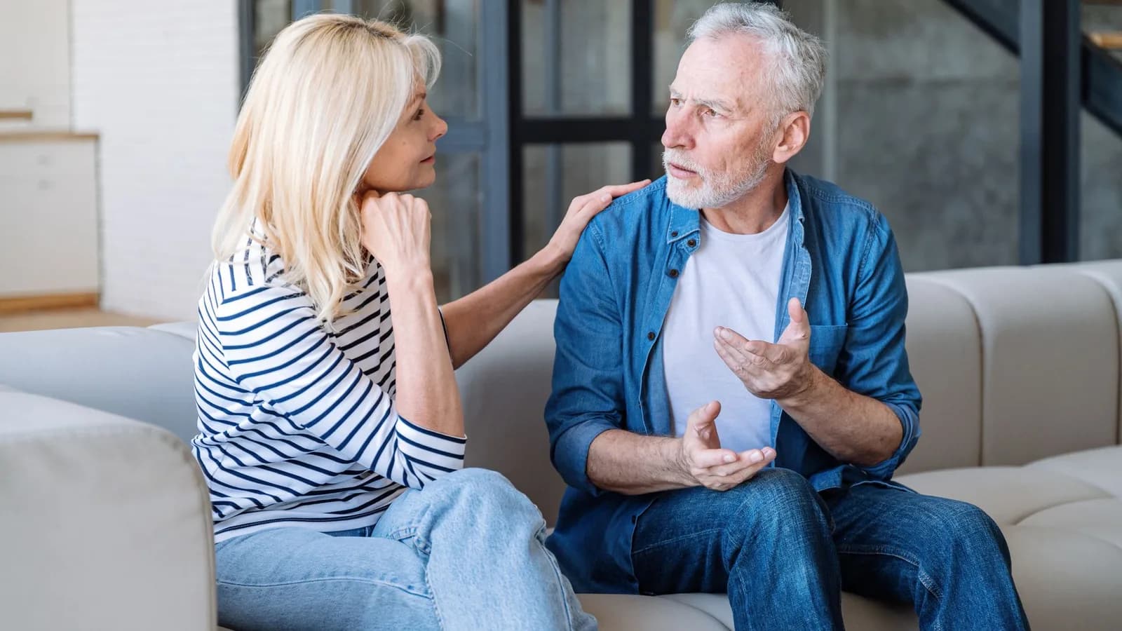 A concerned older man on the couch at home describing a problem to his wife, who rests a hand on his shoulder